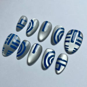 May include: A set of ten silver-colored, oval-shaped false nails. Each nail is decorated with a blue geometric design. The designs vary, including straight lines, curved lines, and abstract shapes. The nails are arranged on a white surface.