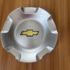 May include: Silver hubcap with a gold and black Chevrolet logo. The hubcap has a circular design with a series of indentations around the edge. The Chevrolet logo is a gold bow tie shape with a black outline.