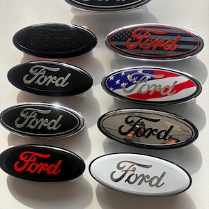 May include: A collection of Ford oval emblems in various designs. The emblems feature the Ford logo in different colors, including black, red, silver, and designs incorporating the American flag. The oval shapes are displayed on a white surface.