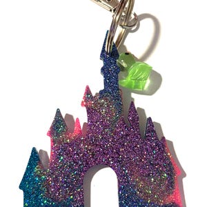 Könnte beinhalten: Ein glitzernder Schlüsselanhänger mit einem Schlossdesign in Blau-, Lila- und Rosatönen. Der Schlüsselanhänger hat einen silbernen Verschluss und einen kleinen grünen Anhänger. Ein verspieltes Accessoire.