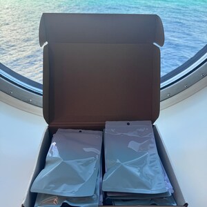 May include: An open cardboard shipping box containing two stacks of white, sealed, opaque pouches. The box has a brown interior and white exterior, with the pouches neatly arranged in two columns inside the container for organized storage.