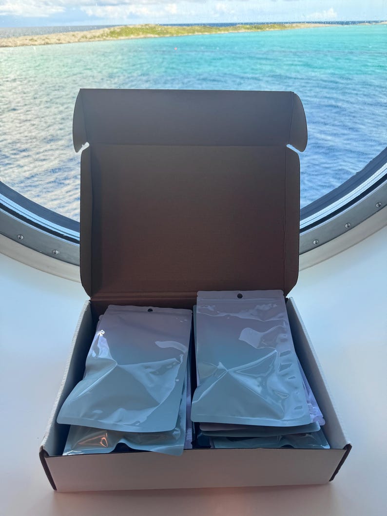 May include: An open cardboard shipping box containing two stacks of white, sealed, opaque pouches. The box has a brown interior and white exterior, with the pouches neatly arranged in two columns inside the container for organized storage.