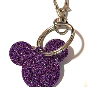 Könnte beinhalten: Ein lila Glitzer-Schlüsselanhänger in Form eines Mäusekopfes. Er hat einen silberfarbenen Karabinerverschluss und einen Schlüsselring zum Befestigen von Schlüsseln. Der Glitzer hat eine tiefviolette Farbe, und der Schlüsselanhänger ist wahrscheinlich aus Acryl oder einem ähnlichen Material.