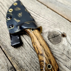 Può includere: Un coltello fatto a mano con un manico in legno e un fodero nero. Il manico presenta un motivo a strisce e il fodero è decorato con dettagli dorati. Il coltello è progettato per l'uso all'aperto.