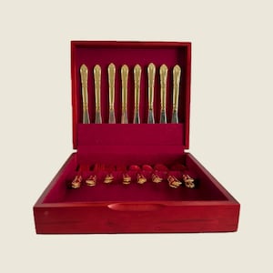 May include: A wooden cutlery case with a red velvet interior holds gold-colored silverware. The set includes knives, forks, and spoons, all with ornate gold handles. The case is open, revealing the arrangement of the utensils.