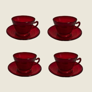 May include: Four ruby red glass teacups and saucers. Each cup has a handle and sits on a matching saucer. The saucers and cups have a textured, ridged design. The set is arranged in a square formation against a neutral background.