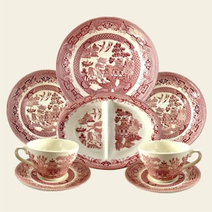 May include: A collection of red and white Willow pattern china. The set includes plates, a divided serving dish, teacups, and saucers. The design features pagodas, bridges, and birds. The dishes are arranged on a light background.