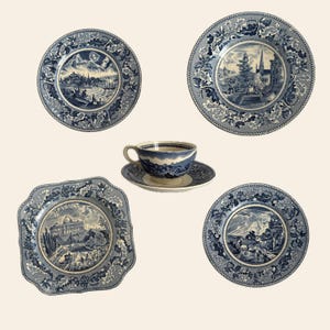 May include: A collection of blue and white ceramic tableware, including four plates and a teacup with saucer. Each piece features a scenic landscape design within a decorative border. The plates are round and square.