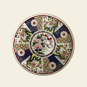 May include: A decorative ceramic plate with a detailed floral design. The plate features a central floral arrangement surrounded by alternating sections of blue, white, and green patterns. The rim is trimmed with gold.