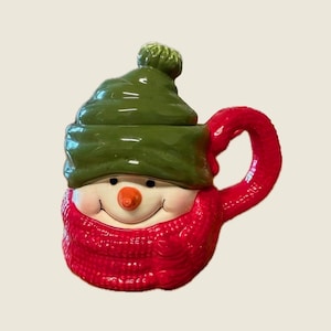 May include: A ceramic snowman-shaped mug with a green hat and a red scarf and handle. The snowman has a carrot nose and a smiling face. A festive holiday-themed mug.