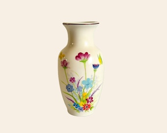 May include: Cream-colored ceramic vase with a floral design. The vase features a bouquet of colorful flowers, including purple, blue, yellow, and red blooms. The rim of the vase is trimmed with a thin brown line.