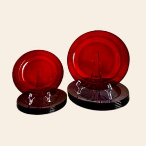 May include: A set of red glass plates, displayed on dark brown plate stands. The plates are round and have a glossy finish. The stands are stacked, and the plates are elevated, showcasing their vibrant color.