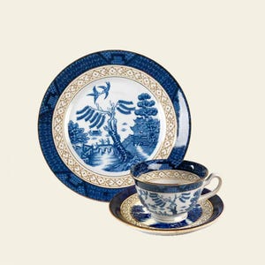May include: A blue and white Willow pattern tea set. The set includes a large plate, a teacup, and a saucer. The plate and cup feature a detailed landscape design with a bridge, trees, and a bird. The edges are trimmed with gold.