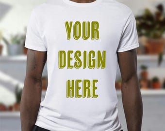 以下が含まれることがあります： オリーブグリーンの太字フォントで「YOUR DESIGN HERE」と書かれた白いTシャツ。半袖のシャツは柔らかい素材で作られているようです。背景はぼやけており、小売店やライフスタイルの設定を示唆しています。