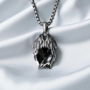 May include: A silver-toned pendant necklace featuring a detailed wing design. The pendant showcases intricate feather detailing and hangs from a matching chain. The necklace is displayed against a soft, white background.