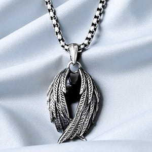May include: A silver-toned necklace with a pendant shaped like stylized wings. The wings are detailed with feather patterns and are crossed. The chain is a linked design. The background is a light blue fabric.
