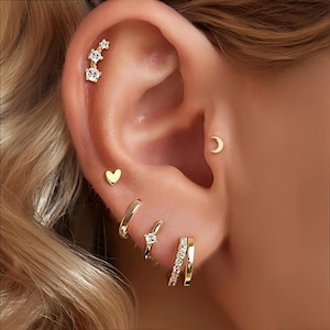 Könnte beinhalten: Ein Ohr mit goldenen Ohrringen. Die Auswahl umfasst einen Herzstecker, einen Halbmondstecker, einen Reifen, einen kleinen Reifen mit einem Edelstein und einen dreifachen Ohrkletterer. Der Hintergrund ist unscharf.