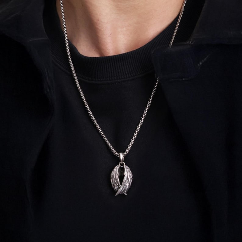 May include: Silver-toned necklace with a pendant featuring a detailed wing design. The pendant is suspended from a delicate chain, creating a striking contrast against the dark background of the person's clothing. The necklace is a stylish accessory.
