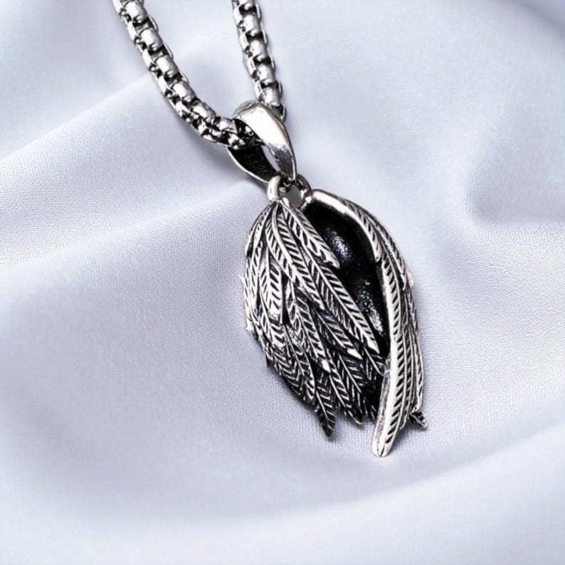 May include: A silver-colored pendant necklace featuring a detailed feather design. The pendant is shaped like a stylized wing, with intricate feather detailing. The chain is a silver-colored, box-style chain. The necklace is displayed on a white, textured background.