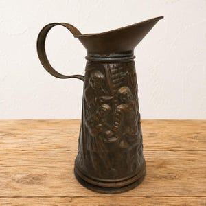 May include: A tall, cylindrical copper pitcher with a wide, flared spout and a curved handle. The exterior surface features a detailed embossed relief depicting a group of figures in a scene, with a dark, aged patina finish across the metal body.