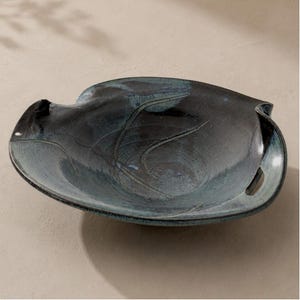 May include: A handcrafted, dark blue ceramic bowl with a unique, organic shape. The bowl features a glossy finish with subtle variations in color, and a textured surface. The design includes a handle-like opening on one side. A decorative piece for the home.