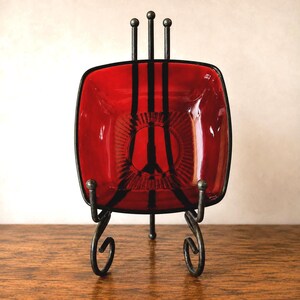 May include: A square red glass bowl with black vertical stripes and a central circular pattern, held upright by a dark metal display stand featuring three vertical prongs at the top and two curved, scroll-like legs at the base.