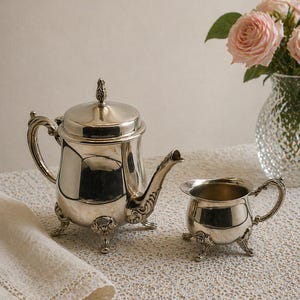 May include: A silver tea set, including a teapot and creamer, sits on a textured surface. The teapot has ornate detailing, a curved spout, and a decorative lid. The creamer has a handle and sits on three small feet. A vase of pink roses is in the background.