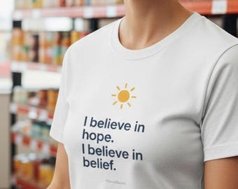 Könnte beinhalten: Weißes T-Shirt mit dem Text "I believe in hope. I believe in belief." und einer gelben Sonne. Die Person trägt das T-Shirt in einem Lebensmittelgeschäft und hält eine Kiste mit Äpfeln.