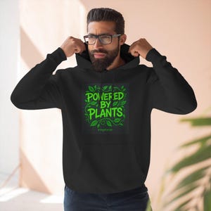 Könnte beinhalten: Schwarzer Kapuzenpullover mit dem Text "POWERED BY PLANTS" in leuchtendem Grün, umgeben von Blättern. Das Wort "Vegetarian" steht darunter. Der Hoodie hat eine Kapuze mit Kordelzug und lange Ärmel.