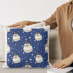 Könnte beinhalten: Quadratisches Zierkissen mit einem sich wiederholenden Muster aus schneemannförmigen Makronen auf dunkelblauem Hintergrund mit weißen Sternen. Die Schneemänner haben schwarze Hüte, Karottennasen und lächelnde Gesichter.