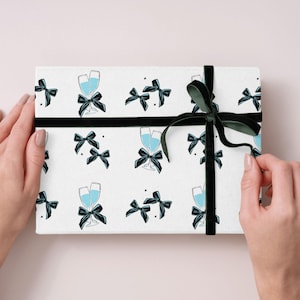 Könnte beinhalten: Eine Geschenkbox mit weißem Hintergrund, verziert mit Champagnergläsern und schwarzen Schleifen. Die Box ist mit einem schwarzen Samtband gebunden. Die Geschenkbox wird von zwei Händen gehalten.