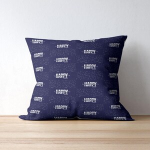 May include: Square throw pillow with a navy blue background and a repeating pattern of white text that reads "HAPPY 2020" and constellations. The pillow is placed on a wooden surface.