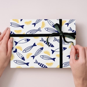 Könnte beinhalten: Ein Geschenkpaket mit einem weißen Hintergrund, der mit marineblauen Fischen und gelben Zitronenscheiben gemustert ist. Es ist mit einem dunkelgrünen Samtband gebunden. Das Geschenk wird von zwei Händen gehalten.