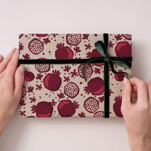 Könnte beinhalten: Ein Geschenk, verpackt mit einem Granatapfelmuster auf beigem Papier, gebunden mit einem dunkelgrünen Samtband. Das Granatapfel-Design umfasst ganze und geschnittene Granatäpfel in Rot- und Marontönen.