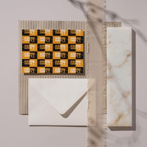 May include: A flat lay featuring a greeting card with a checkered pattern of black and gold squares, each with the text "HAPPY 2026". A white envelope, a textured cardboard piece, and a marble slab are also present.