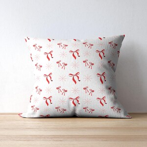 Könnte beinhalten: Quadratisches weißes Kissen mit einem sich wiederholenden Muster aus roten Schleifen und schneeflockenartigen Designs. Das Kissen liegt auf einer Holzoberfläche, der Hintergrund ist eine schlichte weiße Wand.