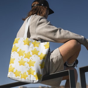 Könnte beinhalten: Weiße Tragetasche mit einem sich wiederholenden Muster aus gelben Cartoon-Sternen mit lächelnden Gesichtern. Die Tasche hat hellblaue Träger. Die Person trägt einen grauen Pullover und eine schwarze Kappe.