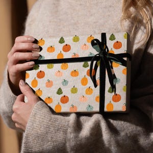 Könnte beinhalten: Eine Geschenkbox, verpackt in Papier mit einem Muster aus orangefarbenen, grünen und rosa Kürbissen auf hellbeigem Hintergrund. Die Box ist mit einem schwarzen Samtband gebunden. Die Box wird von einer Person gehalten, die einen beigen Pullover trägt.