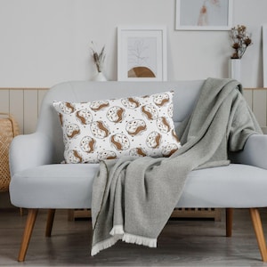 Könnte beinhalten: Ein rechteckiges Kissen mit einem sich wiederholenden Muster aus braunen und weißen Kaffeebohnen-Illustrationen auf weißem Hintergrund. Das Kissen liegt auf einem grauen Sofa, zusammen mit einer passenden Decke.
