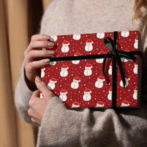 Könnte beinhalten: Ein Geschenk, verpackt mit rotem Papier mit Schneemannmuster und einem schwarzen Band. Das Papier zeigt weiße Schneemänner mit roten Hüten auf rotem Hintergrund mit weißen Punkten. Das Geschenk wird von einer Person gehalten, die einen beigen Pullover trägt.