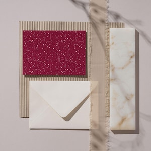 May include: A flat lay featuring a burgundy card with a white constellation pattern, a white envelope, a textured cardboard piece, and a marble-like rectangular block. A beige ribbon adds a decorative touch. The items are arranged on a light gray surface.