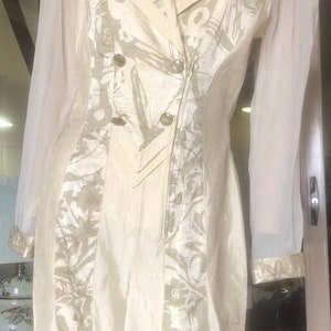May include: A white dress with a double-breasted button closure and a sheer overlay on the sleeves. The dress has a floral pattern and a fitted silhouette.