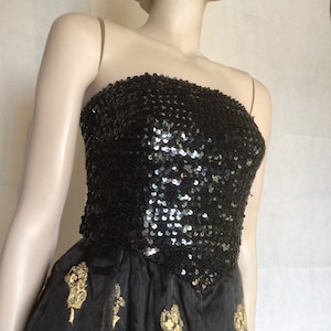 May include: A strapless black dress featuring a bodice covered in shimmering black sequins and a skirt made of black fabric adorned with embroidered gold floral motifs. The garment is displayed on a mannequin, showcasing the contrast between the sequined top and the patterned skirt.