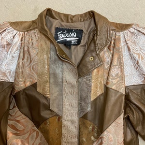 May include: A brown leather jacket with a patchwork design featuring different textures and colours, including gold, white, and brown. The jacket has a collar and a button closure. The label on the jacket reads "Fantasia".