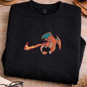 May include: Black crewneck sweatshirt featuring an embroidered orange and red dragon with teal wings and a fiery tail. The dragon is in a dynamic pose, with flames emanating from its mouth. The sweatshirt is made of a soft material.
