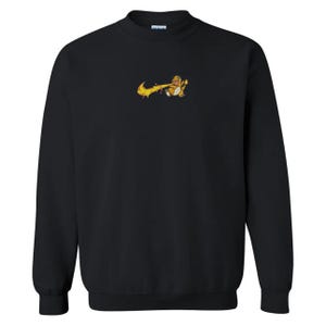 May include: Black crewneck sweatshirt with a small, embroidered design of a cartoon character breathing fire. The character is yellow and orange, with a flame-like design. The sweatshirt is a solid black color.