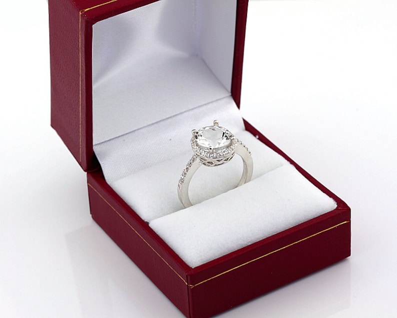 May include: A silver ring with a large, clear, round gemstone surrounded by a halo of smaller gemstones. The ring is in a red velvet ring box.