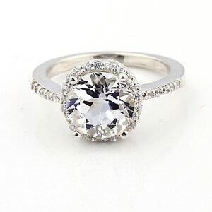 May include: A silver ring with a large, clear, round gemstone surrounded by a halo of smaller gemstones. The band is also set with smaller gemstones.