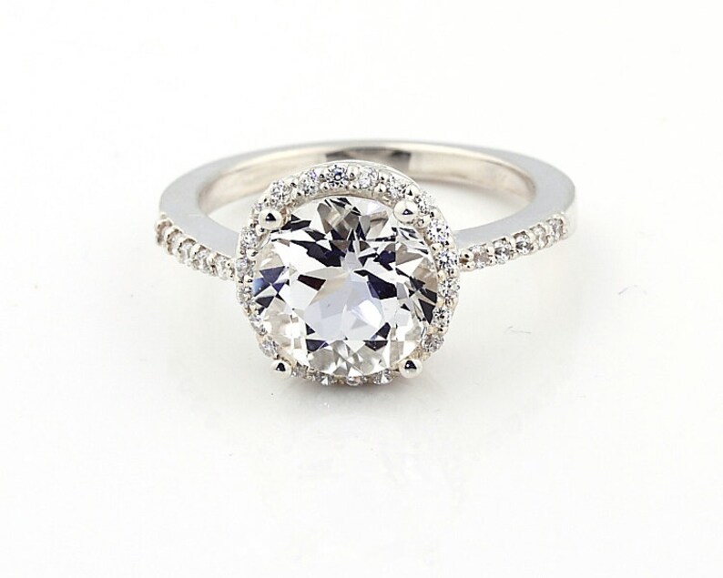 May include: A silver ring with a large, clear, round gemstone surrounded by a halo of smaller gemstones. The band is also set with smaller gemstones.