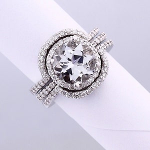 May include: A white gold engagement ring with a large, round, clear center stone surrounded by a halo of smaller diamonds. The ring is set with a double band of smaller diamonds.